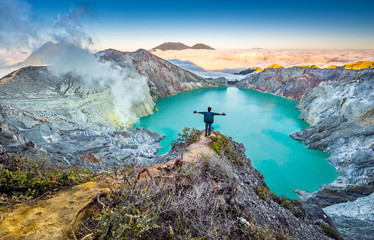 Man hiker staying on crater cliff with spread his arms. Panorama of colourful sunrise daybreak on the edge of volcano over acid lake in Kawah Ijen crater, Java, Indonesia. Smoke from sulphur mining.