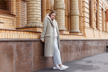 Wall Mural - Beautiful young girl in fashionable biege hoodie, elegant gray coat and pants on brick wall background. Walking on the street.