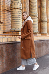 Wall Mural - Beautiful young girl in fashionable biege hoodie, elegant brown camel coat and pants on brick wall background. Walking on the street.