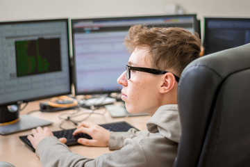  Male portrait in profile. Teenager looks at the monitor screens. Side view. A young guy with glasses sits in a chair in front of several monitors. The programmer works in the office.