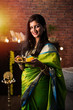 Indian Girl / women holding Pooja Thali while wearing green saree and sitting at home with flower rangoli and Samai, celebrating Diwali or hindu festival. selective focus