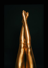 Beautiful And Slendel Female Legs Covered With Golden Paint, Posing On Black Background, Beauty And Fashion Concept