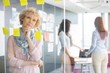 © MDBPIXS - Thoughtful businesswoman reading sticky notes on glass with colleagues in background