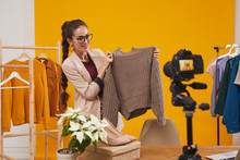 Portrait Of Contemporary Young Woman Holding Knit Sweater And Smiling At Camera While Filming Video For Fashion And Beauty Channel Against Yellow Background, Copy Space
