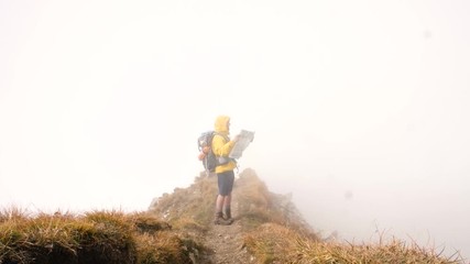 Wall Mural - young man hiker on mountain trail reading map. cloudy sky and fog. yellow hood jacket and backpack. 