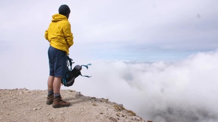 Wall Mural - young man hiker standing on mountain peak with cloudy sky and fog. yellow jacket, backpack and beani