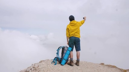 Wall Mural - young man hiker standing on mountain peak taking pictures with cloudy sky and fog. yellow jacket, ba