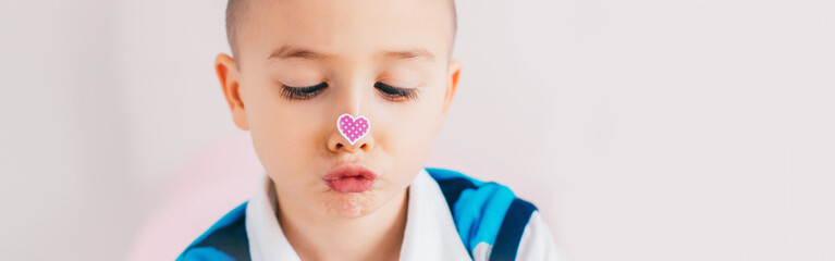 Wall Mural - Cross-eyed kid looking at purple violet heart sticker on his nose. Funny hilarious white Caucasian cute adorable child boy making faces. Valentine Day holiday concept. Web banner header for website.