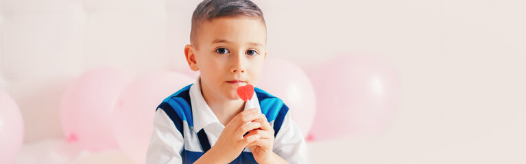 Wall Mural - Happy Caucasian pensive child boy eating heart shape red lollipop candy. Valentine Day love holiday concept. Web banner header for website.