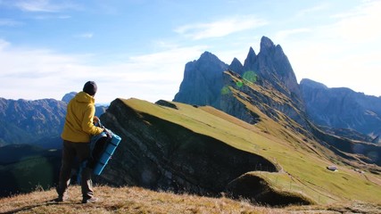 Wall Mural - young man hiking at seceda mountain taking off backpack on the peak at sunrise. ellow jacket, boots,