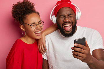 Poster - Emotive laughing teenage ethnic female and male watch funny video on modern device, express positive emotions and feelings, cannot stop giggling, stands next to each other against pink background