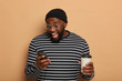 Portrait of overjoyed dark skinned guy concentrated in smartphone, gets nice message, expresses happy surprisement, wears big spectacles, striped sweater, drinks takeaway coffee, isolated on beige