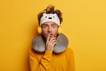 Hipster male passenger stares with bugged eyes, enjoys flight in first class, listens music via headphones on board, wears travel pillow, prepares for sleep in transport, isolated on yellow wall