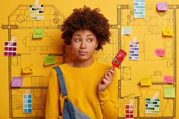 Thoughtful ethnic woman stands with paint brush, dressed in yellow jumper and overalls, poses against room plan with stickers and color palette, repairs house, thinks about decor, busy refurbish wall