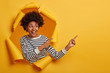 © wayhome.studio  - Studio shot of attractive curly girl шn striped jumper points aside on blank space with both index fingers, stands in paper hole of yellow background, happy to show cool promotion or advert.