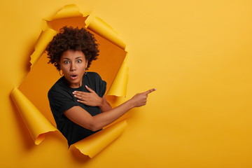 Wall Mural - Stunned Afro American woman looks with scared speechless expression, holds breath from excitement, demonstrates something on right side, stands in paper hole, wears black t shirt advertises copy space