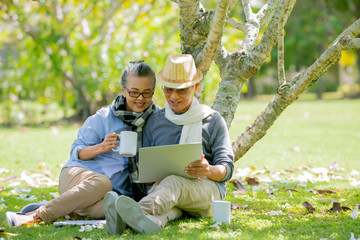 Wall Mural - Relax Senior Asian couple  sitting under the tree  at park  using  laptop computer while holding coffee cup