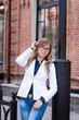 © Марина Демешко - A cute girl in the white jacket with glasses  is adjusting her hair and looking sideways with shy smile while standing outdoors on the background of a brick wall. Dressed in business style