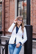 © Марина Демешко - A pretty girl in a white jacket with glasses looking sideways with beautiful smile while standing outdoors on the background of a brick wall and flirting with guy she likes. Dressed in business style