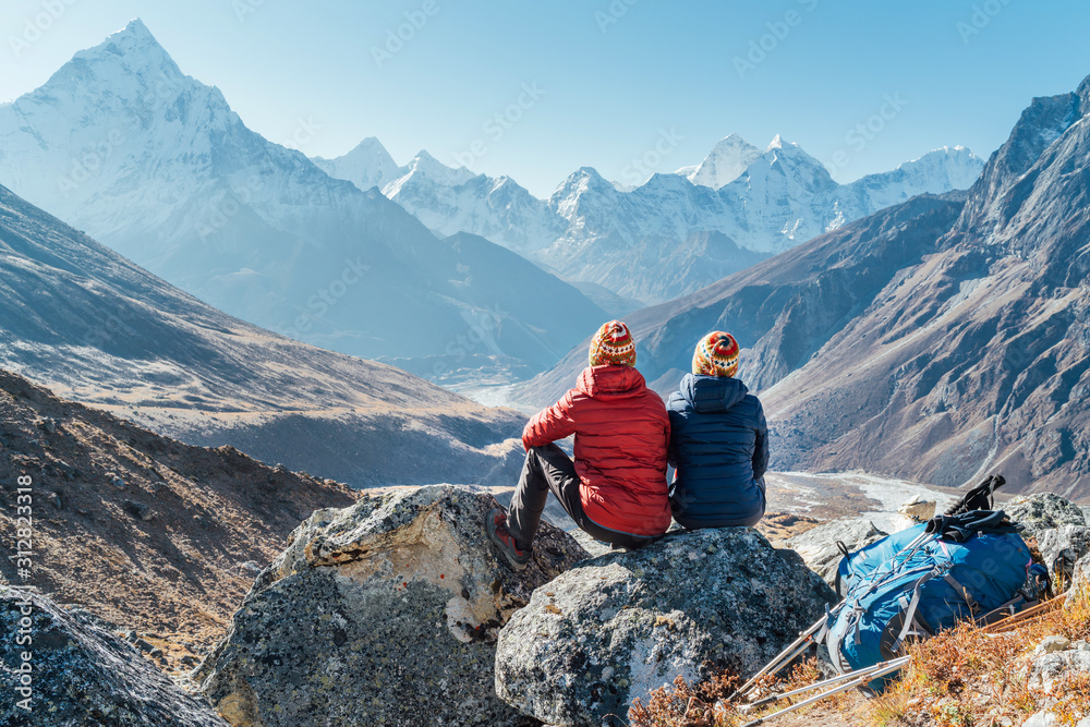 Couple resting on the Everest Base Camp trekking route near Dughla 4620m. Backpackers left Backpacks and trekking poles and enjoying valley view with Ama Dablam 6812m peak and Tobuche 6495m - obrazy, fototapety, plakaty 