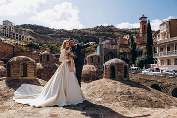 Wall Mural - Stylish couple of brides: Georgian-style wedding, man with mustache dressed in costume and pretty blonde bride in wedding dress against beautiful city background