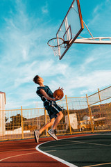 Sports and basketball. A young teenager in a blue tracksuit throws a ball into the basket in a powerful jump. Blue sky in the background