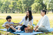 Asian Family Having Picnic In Fresh Green Park Together. Mother Touching To Kid With Smile While Grandmother Looking At Child Playing Ukelele Or Mini Guitar With Happiness. Relax Family Activity.