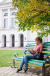 © Марина Демешко - A beautiful female student in a polka dot shirt is sitting under a tree on a green bench and reading a book on the background of a university. Sunny autumn day