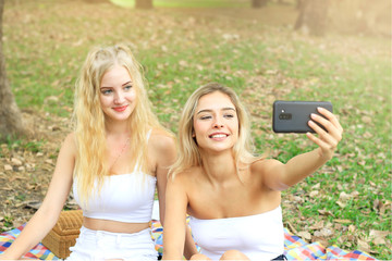 Wall Mural - Happy friends on picnic in park, two young beautiful teen girls taking selfie with smartphone and having fun while spending time together at summer garden, woman outdoor friendship concept.