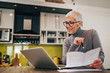 © bnenin - Happy woman looking at laptop, holding documents and sitting in the kitchen, portrait.