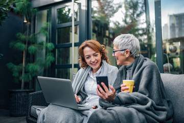 Wall Mural - Cheerful mother and daughter sitting at the backyard, using laptop and looking at smart phone together, portrait.