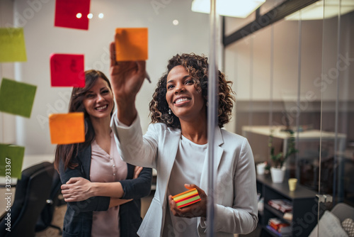 Fotografía Creative businesswoman writing on sticky notes on a glass wall, female colleague looking