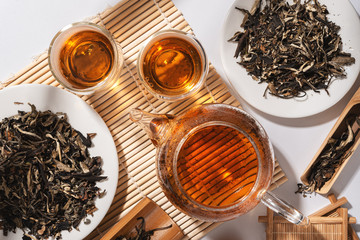 Wall Mural - A teapot with freshly brewed tea and glass cups with hot drinks, Dry green tea leaves on a white plate, bamboo accessories for the brewing ceremony, shade from trees on the background