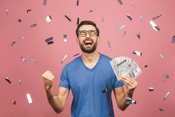 Happy winner! Young rich man in casual t-shirt holding money dollar bills with surprise isolated over pink background.