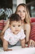 © Dana Keli - a beautiful young blonde smiling woman with long hair in a dress lies on a sofa with pillows. Her cute daughter toddler creeps forward on camera. Mom in defocus in the background. multiracial family