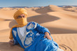 Bedouin nomad, Sahara desert, Morocco. Portrait of a Bedouin nomad with colorful turban and big smile sitting on sand dune popular tourist spot. A tuareg man portrait with his traditional clothes.
