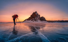 Photographer Wear Red Clothes Take Photo Of Shamanka Rock At Sunrise With Natural Breaking Ice In Frozen Water On Lake Baikal, Siberia, Russia.