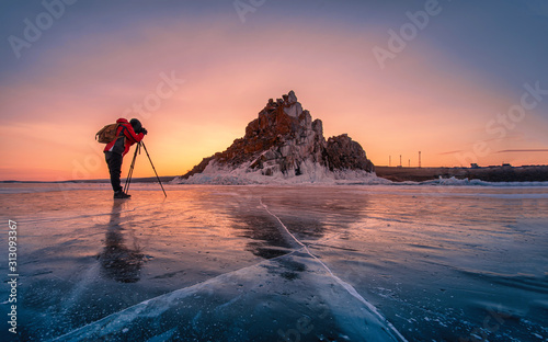 Photographer wear red clothes take photo of Shamanka rock at sunrise with natural breaking ice in frozen water on Lake Baikal, Siberia, Russia.