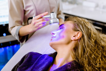 Wall Mural - Skin Cosmetology. Woman beautician Doing Blue Light Therapy On face of pretty young blond woman