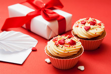 Close-up Of Two Cupcakes With Cream And Heart Decor On A Red Background With Gift And Envelope.