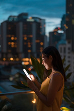 Smiling Woman Standing On Roof Terrace At Dusk Looking At Cell Phone