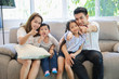 © feeling lucky - Asian father, mother, older little young daughter and son child sitting on sofa in living room watching their favourite TV show at home and having fun together. Young happy family portrait concept.