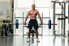 A Strong Muscular Shirtless Mature Older Bodybuilding Athlete With Balding Gray Hair  Holding A Heavy Barbell, Looking At Camera In A Gym