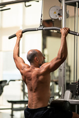 Wall Mural - Muscular shirtless mature older bodybuilding athlete with balding gray hair smiling at the camera performing lat pulldown exercise on a machine in a gym