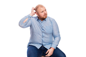 A fat red-haired man with a beard in a blue shirt and jeans sits and scratches his head. Isolated over white background.