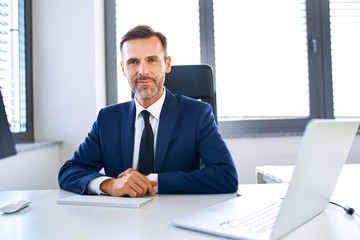 Wall Mural - Smiling mature businessman sitting at office desk looking at camera