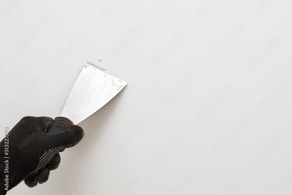 Removing old paint from the wall with a metal spatula. Light background. Working tool, spatula in hand in black gloves against the background of a white wall, work plasterer. Copy space. - obrazy, fototapety, plakaty 