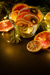 Wall Mural - Creative moody holiday Christmas New Year food fruits with dried grapefruit, kiwi, orange and lemon in glass bowl with branch of fir tree with warm led lights, angle view, copy space