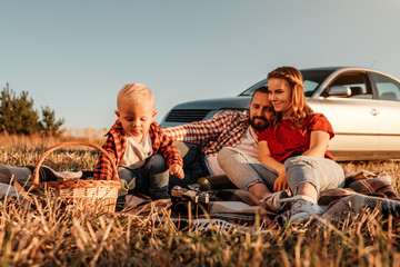 Happy Young Family Mom and Dad with Their Little Son Enjoying Summer Weekend Picnic Sitting on the Plaid Near the Car Outside the City in the Field at Sunny Day Sunset, Vacation and Road Trip Concept