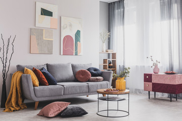 Wall Mural - Real photo of abstract paintings hanging on white wall above a gray sofa in a living room interior with big windows